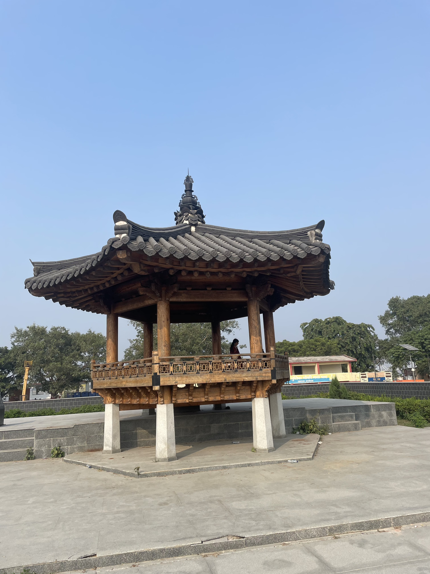 A traditional wooden pavilion with a curved, tiled roof stands on a stone platform against a clear blue sky.
