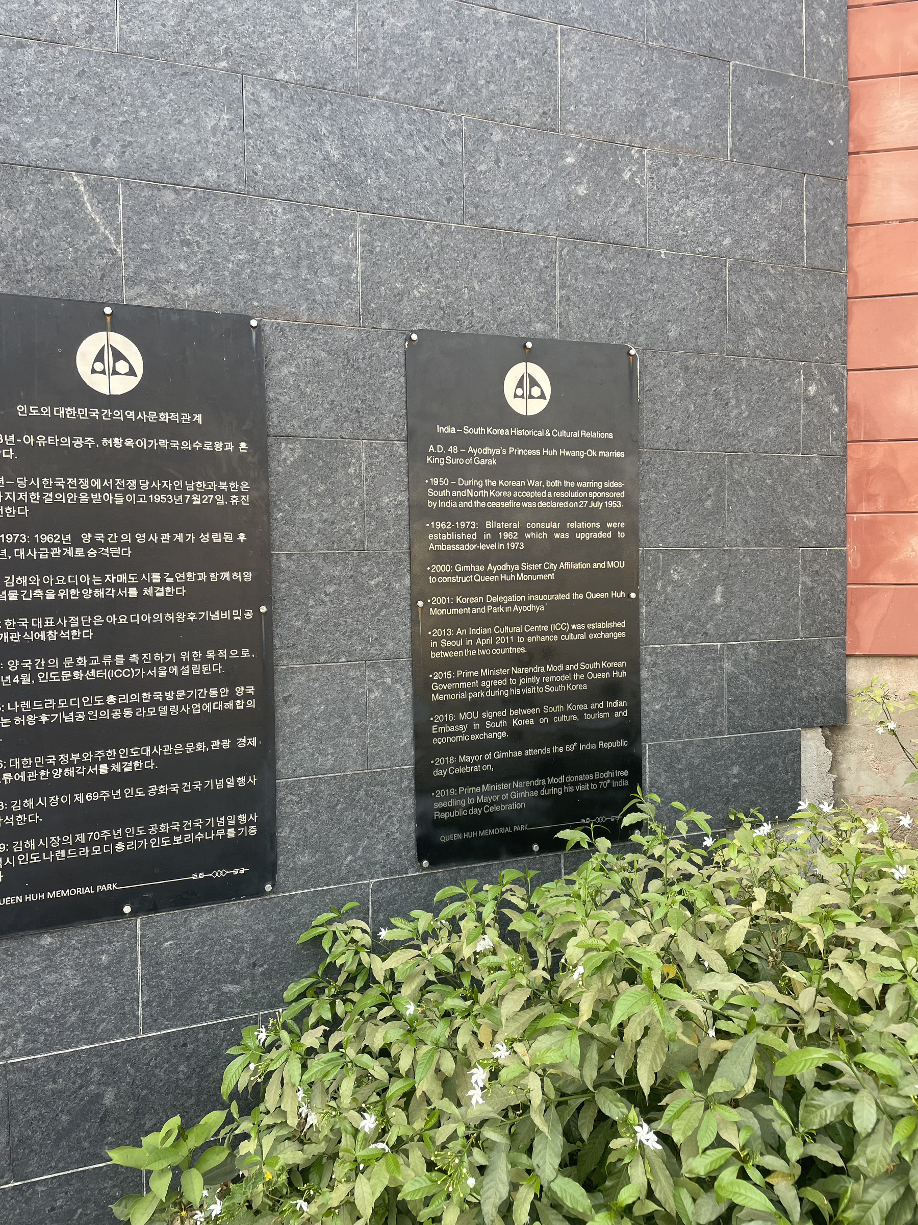 Two plaques with text in Korean and English are mounted on a stone wall above a row of green plants.